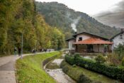 ljudi šeću stazom, lijevo šuma, desno potok, pored potoka kuća iz čijeg dimnjaka se dimi