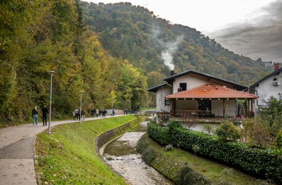 ljudi šeću stazom, lijevo šuma, desno potok, pored potoka kuća iz čijeg dimnjaka se dimi