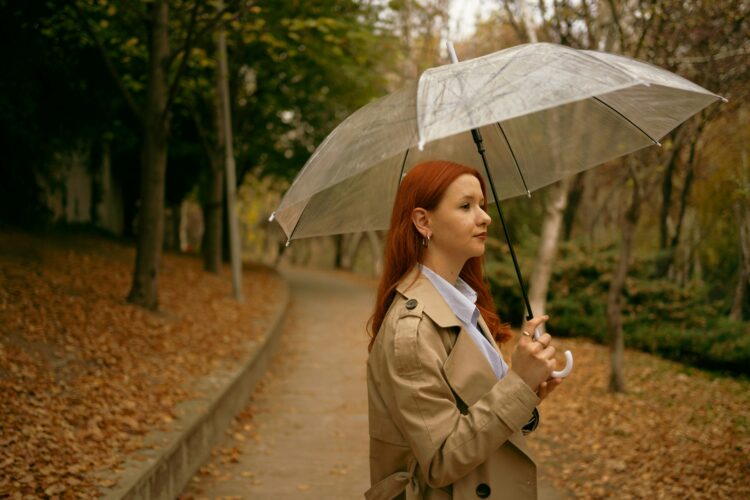 Žena s riđom kosom drži kišobran u ruci te nosi kaput i stoji na cestici u parku za vrijeme jeseni