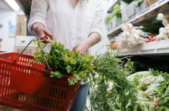 zeleno lisnato povrće viri iz crvene košare u trgovini koju nosi žena u bijeloj košulji