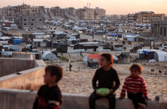 troje djece sjede na zidiću, iza njih gaza